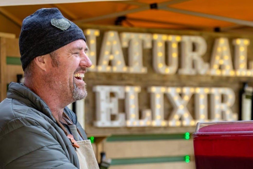 Brett Austin laughing beside the illuminated Natural Elixir sign at his market stall